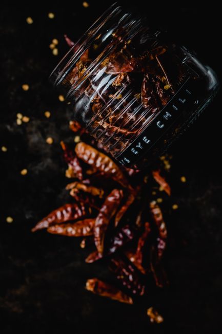 Ancho - Overturned glass jar spilling dried red chili peppers on a dark surface, highlighting spicy flavor.